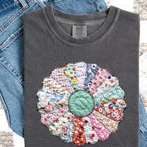 May include: A gray Comfort Colors t-shirt with a colorful, patchwork flower design. The flower features various patterned fabrics in shades of red, blue, pink, and green. The shirt is folded on top of a pair of blue jeans.