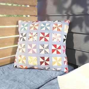 May include: Square throw pillow with a patchwork design. The pillow features a light blue background with a grid of colorful pinwheel patterns in red, white, blue, and yellow. The pillow is placed on a blue cushion.
