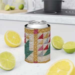 May include: A can cooler with a quilted design. The cooler features a patchwork pattern with red, green, and blue triangles and squares on a cream background. The edges are trimmed with a gold-colored fabric. The cooler is designed to hold a standard-sized can.