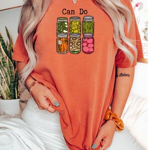 Puede incluir: Una camiseta de manga corta color naranja quemado con un estampado gr&aacute;fico de seis frascos de vidrio llenos de varios vegetales encurtidos y el texto "Can Do" impreso en negro sobre los frascos. Los frascos contienen zanahorias, jud&iacute;as y r&aacute;banos.