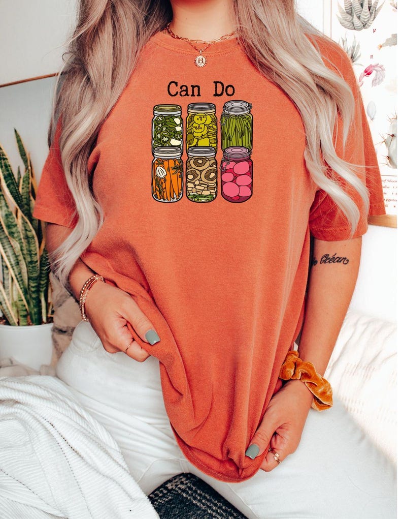 Puede incluir: Una camiseta de manga corta color naranja quemado con un estampado gr&aacute;fico de seis frascos de vidrio llenos de varios vegetales encurtidos y el texto "Can Do" impreso en negro sobre los frascos. Los frascos contienen zanahorias, jud&iacute;as y r&aacute;banos.