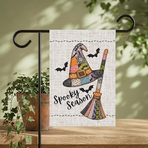 May include: A rectangular garden flag with a beige textured background featuring a patchwork witch hat and broomstick illustration. The design includes black bats and the text Spooky Season in a cursive font. The flag hangs on a black metal stand.