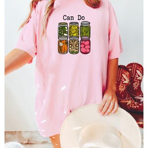 Puede incluir: Una camiseta de manga corta de color rosa claro con un estampado gr&aacute;fico de seis frascos de vidrio llenos de diversas verduras encurtidas y el texto "Can Do" impreso en negro sobre ellos. El dise&ntilde;o incluye frascos de pimientos, pepinillos, jud&iacute;as verdes, zanahorias, champi&ntilde;ones y r&aacute;banos.