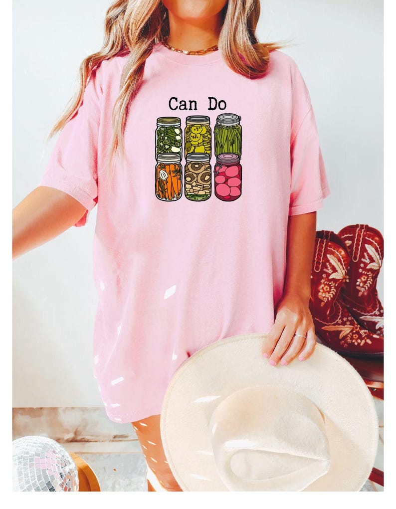 Puede incluir: Una camiseta de manga corta de color rosa claro con un estampado gr&aacute;fico de seis frascos de vidrio llenos de diversas verduras encurtidas y el texto "Can Do" impreso en negro sobre ellos. El dise&ntilde;o incluye frascos de pimientos, pepinillos, jud&iacute;as verdes, zanahorias, champi&ntilde;ones y r&aacute;banos.