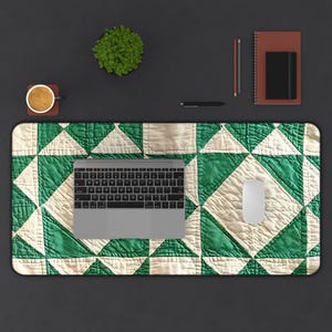 May include: A green and cream-colored quilted desk mat with a laptop, mouse, and other office supplies. The mat features a geometric pattern of green and cream-colored squares and triangles. A small plant, a cup of coffee, a notebook, and a pen are also on the desk.