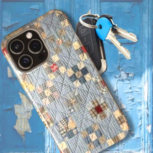 May include: A smartphone case featuring a quilted fabric pattern with light blue, tan, and red checkered squares, positioned next to a black car key fob with a bright blue plastic key cap and two silver metal keys attached to a silver ring.