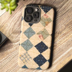 May include: A phone case with a quilted design featuring a patchwork pattern in shades of blue, cream, and floral accents. The case has a textured appearance and a camera cutout.