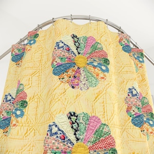 May include: A yellow shower curtain featuring a quilted texture and a repeating pattern of large, multi-colored floral motifs. Each flower consists of various patterned fabric petals in shades of pink, blue, green, and red, centered with a solid yellow circle, all suspended from a curved metal rod with silver rings.
