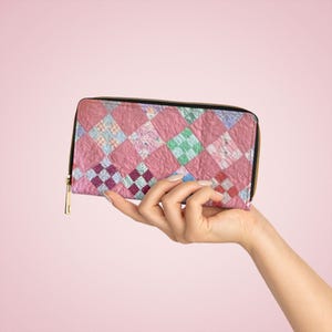 May include: A pink quilted pattern wallet with a black zipper and gold-colored zipper pull. The wallet features a patchwork design with various shades of pink, purple, and green squares. The wallet is held up in the air.