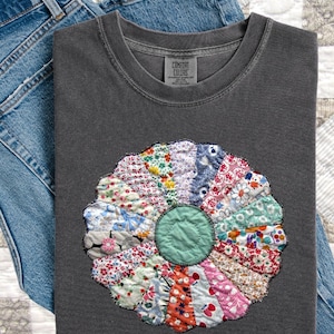 May include: A gray Comfort Colors t-shirt with a colorful, patchwork flower design. The flower has a green center and petals in various patterned fabrics. The shirt is folded on top of blue denim jeans.