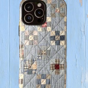 Puede incluir: Una funda para smartphone con un estampado de patchwork acolchado en tonos azul claro, crema y azul marino. El dise&ntilde;o muestra una cuadr&iacute;cula de bloques cuadrados con diversas texturas de cuadros y rayas, acentuados por peque&ntilde;os cuadrados rojos y tostados.