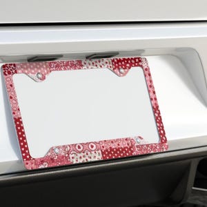 May include: A decorative license plate frame with a patchwork design in shades of red and pink. The frame features various patterns, including hearts and polka dots, and is mounted on a white vehicle.