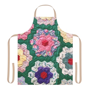 May include: A floral patterned apron with a quilted design. The apron features a patchwork of hexagonal shapes in various colors, including pink, red, purple, yellow, and green. The apron has tan straps and a neck loop.