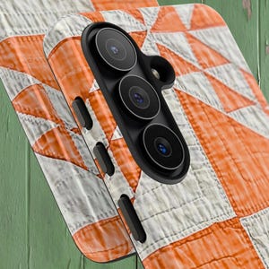 May include: A smartphone case featuring a white and orange geometric quilt pattern. The case has a black camera module housing three circular lenses and a flash, along with black side buttons integrated into the frame of the protective cover.