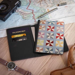 May include: A black passport holder featuring a front cover with a multi-colored pinwheel quilt pattern. The interior includes slots for a passport, a gold-colored credit card, and a black credit card. The passport cover is open and displayed flat.