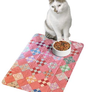 May include: A white cat with gray markings sits above a pink quilted pet mat with a white bowl filled with pet food. The mat features a patchwork design with various patterned squares in shades of pink, red, and other colors.