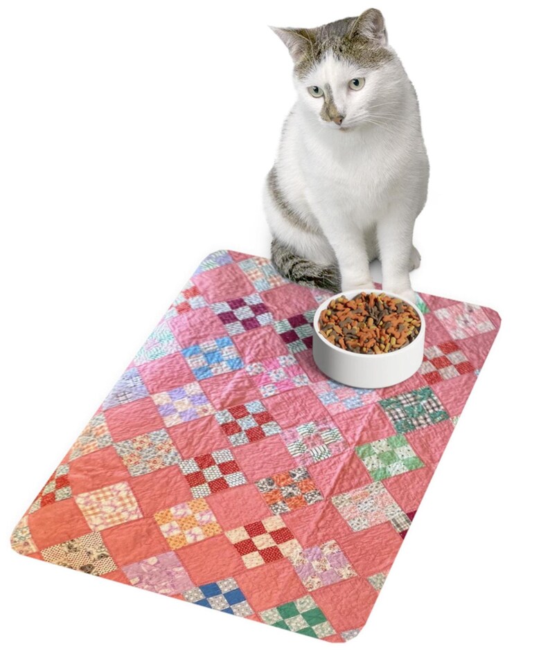 May include: A white cat with gray markings sits above a pink quilted pet mat with a white bowl filled with pet food. The mat features a patchwork design with various patterned squares in shades of pink, red, and other colors.