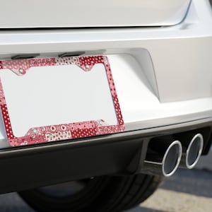 May include: A decorative license plate frame with a patchwork design in shades of red and pink. The frame is mounted on the back of a white car, near the exhaust pipes. The pattern includes polka dots, hearts, and floral motifs.