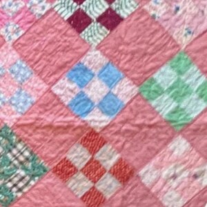 May include: Close-up of a vintage quilt with a patchwork design. The quilt features a repeating pattern of diamond shapes in various colors, including pink, blue, green, red, and white. The quilt has a soft, textured appearance, suggesting it is made of fabric.