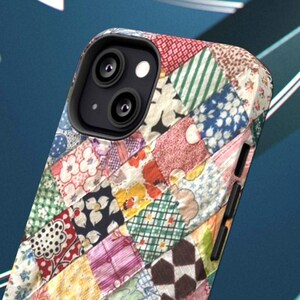 Op de afbeelding: Een smartphonehoesje met een veelkleurig patchwork-quiltpatroon met diverse bloemmotieven, geometrische vormen en ruitjes in tinten rood, blauw, groen, geel en wit, voorzien van zwarte zijknoppen en een zwarte omlijsting voor de cameralens.