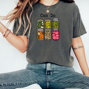Puede incluir: Una camiseta gris carb&oacute;n de manga corta con un gr&aacute;fico de seis frascos de vidrio llenos de varios vegetales encurtidos y el texto Can Do impreso en negro sobre ellos. Los frascos contienen pimientos verdes, pepinillos, jud&iacute;as verdes, zanahorias, champi&ntilde;ones y r&aacute;banos rosas.