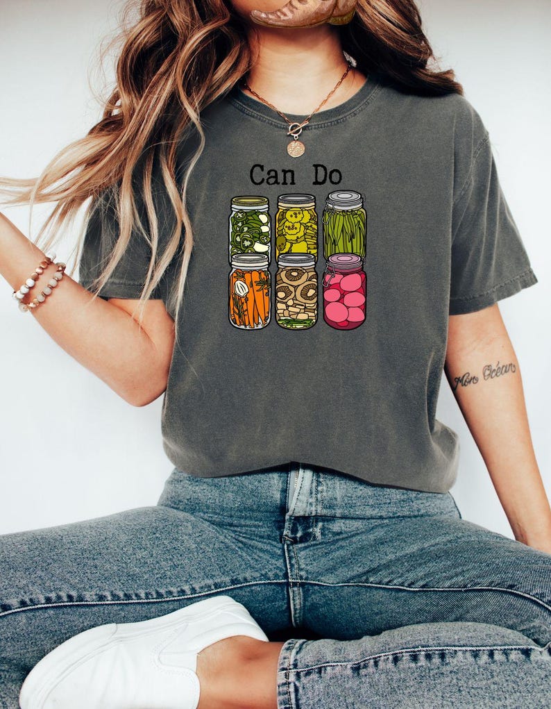 Puede incluir: Una camiseta gris carb&oacute;n de manga corta con un gr&aacute;fico de seis frascos de vidrio llenos de varios vegetales encurtidos y el texto Can Do impreso en negro sobre ellos. Los frascos contienen pimientos verdes, pepinillos, jud&iacute;as verdes, zanahorias, champi&ntilde;ones y r&aacute;banos rosas.