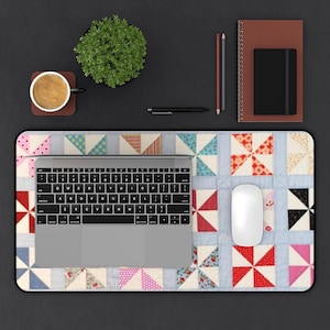 May include: A desk setup featuring a laptop, mouse, and a patterned mousepad with a quilt design. The quilt pattern includes red, blue, pink, and white pinwheel blocks. A cup of coffee, a small plant, and stationery are also on the dark surface.