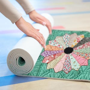 May include: A white yoga mat being rolled up next to a green quilted mat with a colorful, patchwork flower design. The flower has a black center and petals in various patterned fabrics. The mats are on a light-colored surface.