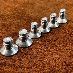 May include: Six silver metal screws with wide, flat heads are arranged diagonally on a textured brown surface. The screws have a threaded shaft and a circular top. The heads are slightly angled.