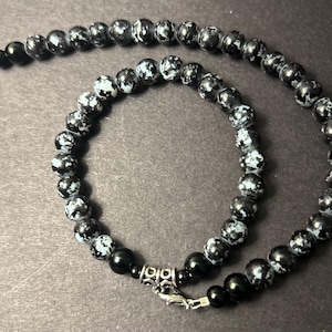 May include: A black beaded necklace with a mix of solid and speckled beads. The necklace features a silver clasp and connector. The beads have a mix of black and light blue speckles.