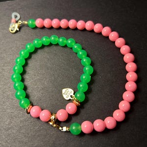 May include: A beaded eyeglass chain and bracelet set. The chain features pink and green beads with gold-colored clasps. The bracelet is made of green beads and has a small heart-shaped charm that says "Made with Love".
