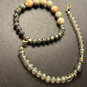 May include: A beaded necklace and bracelet set. The bracelet features faceted, dark gray beads and a mix of round, patterned stone beads in shades of orange, gray, and cream. The necklace has clear, faceted beads and gold-toned accents.
