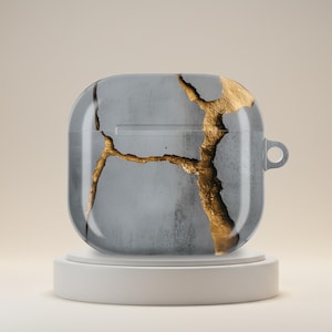 May include: Clear protective case for wireless earbuds with a gray background and gold-colored crack pattern. The case has a loop for attaching to a keychain or lanyard. The case is displayed on a white pedestal.