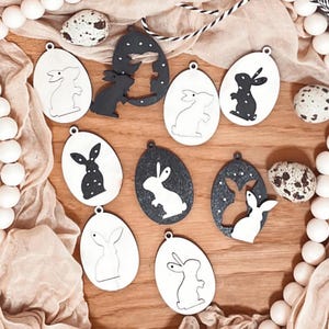 Puede incluir: Nueve adornos de huevos de Pascua de madera con siluetas de conejos. Los adornos están pintados en blanco y negro y tienen un acabado moteado. Algunos de los conejos están recortados, mientras que otros son sólidos.