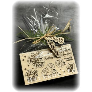 May include: A wooden gift box with a clear plastic wrap. The box has a variety of engraved designs, including a sun, a rocking horse, a baby carriage, a pacifier, and footprints. The box also has the text "Hallo Sonne vom Bauch mitten ins Herz" and "Katharina ich bin da!" The box is tied with a brown ribbon and a tag that says "Zur Taufe Katharina".