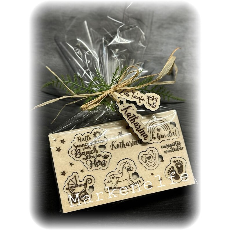 May include: A wooden gift box with a clear plastic wrap. The box has a variety of engraved designs, including a sun, a rocking horse, a baby carriage, a pacifier, and footprints. The box also has the text "Hallo Sonne vom Bauch mitten ins Herz" and "Katharina ich bin da!" The box is tied with a brown ribbon and a tag that says "Zur Taufe Katharina".