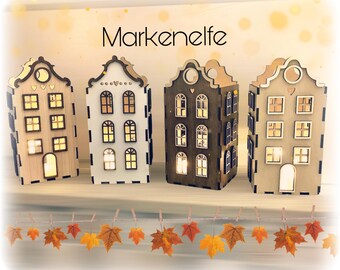 May include: Four wooden houses with lit windows, each with a different color scheme. The houses are arranged in a row on a shelf with a garland of fall leaves hanging below. The text "Markenelfe" is displayed above the houses.