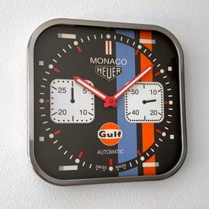 Könnte beinhalten: Quadratische Wanduhr mit schwarzem Zifferblatt, weißen quadratischen Zifferblättern und roten Zeigern. Die Uhr hat blaue und orangefarbene Streifen, das "Gulf"-Logo und den Text "MONACO HEUER". Die Uhr ist in einem silbernen Metallrahmen eingefasst.