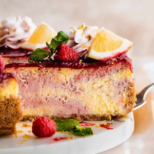 May include: A slice of lemon raspberry cheesecake with a pink and yellow swirl, topped with a lemon wedge, raspberries, and mint leaves. The cheesecake is on a white cake stand with a bowl of raspberries in the foreground.