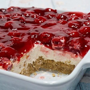May include: A white baking dish filled with a cherry dessert. The dessert has a layer of graham cracker crumbs, a layer of cream cheese, and a layer of cherry topping. A portion of the dessert is missing, revealing the layers.