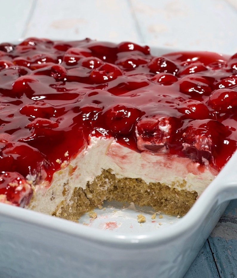 May include: A white baking dish filled with a cherry dessert. The dessert has a layer of graham cracker crumbs, a layer of cream cheese, and a layer of cherry topping. A portion of the dessert is missing, revealing the layers.