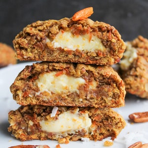 Op de afbeelding: Een stapel van drie wortelcakekoekjes, doorgesneden om een romige witte vulling te onthullen. De koekjes zijn gevuld met wortelen en noten, met een stukje wortel bovenop. Pecannoten en kokosvlokken zijn verspreid.