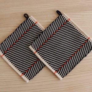 May include: Two black and white woven pot holders with orange accents. The pot holders have a diamond pattern.