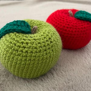 Könnte beinhalten: Zwei gehäkelte Äpfel, einer grün und einer rot, liegen nebeneinander. Jeder Apfel hat ein grünes Blatt und einen braunen Stiel. Die Äpfel sind handgefertigt und wirken weich und plüschig.