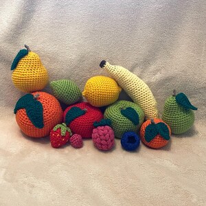 Könnte beinhalten: Eine Sammlung gehäkelter Früchte und Gemüse, darunter eine Birne, eine Zitrone, eine Banane, eine Orange, ein Apfel, eine Erdbeere, eine Blaubeere und ein grünes Gemüse. Die gehäkelten Artikel sind alle in leuchtenden Farben gehalten und haben realistische Details.