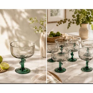 May include: Clear glass coupe glasses with a green cactus-shaped stem. The glasses are arranged on a marble surface, with a few limes and decorative plants in the background. The glasses are designed for cocktails.