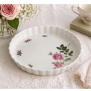 Może przedstawiać: Białe ceramiczne naczynie do ciasta z falistym brzegiem, ozdobione ilustracjami różowych róż i zielonych liści. Okrągłe naczynie stoi na białym koronkowej obrusie. W tle wazon z różowymi różami i filiżanka do herbaty.