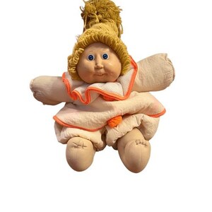 May include: A vintage doll with a soft, fabric body and a vinyl face. The doll has blue eyes, a yellow yarn wig, and is wearing a white dress with orange trim. The doll's arms and legs are also made of fabric.