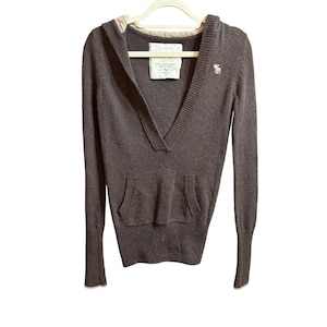 May include: A dark brown hooded sweater with a V-neck and a kangaroo pocket. The sweater has long sleeves and a small logo on the chest. The hood and cuffs have a light-colored trim.