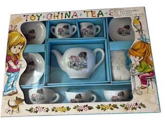 May include: A vintage toy china tea set in a blue box. The set includes a teapot, cups, saucers, and a serving plate, all decorated with a child illustration. The box has the text "TOY CHINA TEA" and illustrations of children.